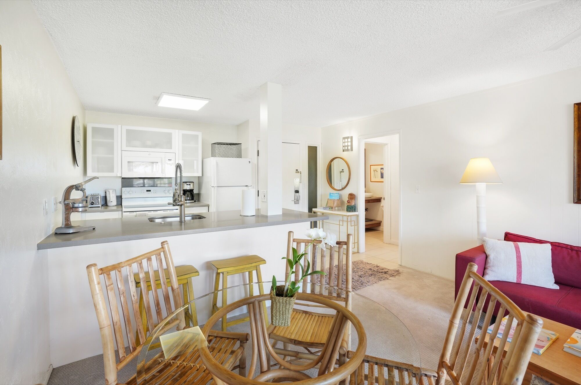 A modern kitchen with white cabinets, a built-in microwave, and a refrigerator. There is a dining area with a round table and chairs, and a red sofa in the living room.