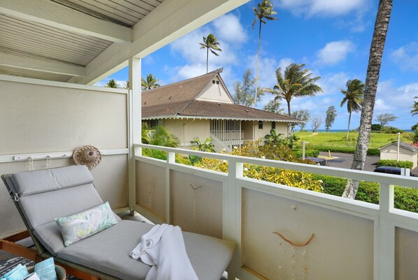 A balcony with a lounge chair, a small table, and a view of a house with a tiled roof and palm trees. - Beach, shops, restaurants are steps away! (Kapaa)