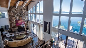 A spacious living area with a stone fireplace, large windows offering a view of the water, and a high ceiling with wooden beams. -  Beautiful Lakefront Home with Pier and Buoy. (Glenbrook)
