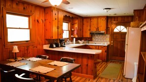 Interior de una cabaña de madera con una mesa de comedor, sillas, una cocina con refrigerador y una ventana.