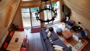 A cozy cabin interior with a wooden floor, a dark sofa with cushions, a dining table with red chairs, and a large window offering a view of the outdoors. - A-Frame Cabin Escape in George Washington Natl Forest, Lost River Area, WV (Wardensville)