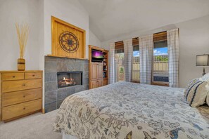 Une chambre avec une cheminée, un lit recouvert d’une couvre-lit à motifs floraux, une commode en bois et une vue sur l’extérieur.