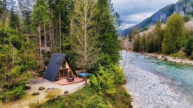 A cabin with a sloped roof, surrounded by dense forest and a river.