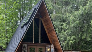 A triangular wooden cabin with a stone entrance, a patio with outdoor furniture, and a surrounding forest.