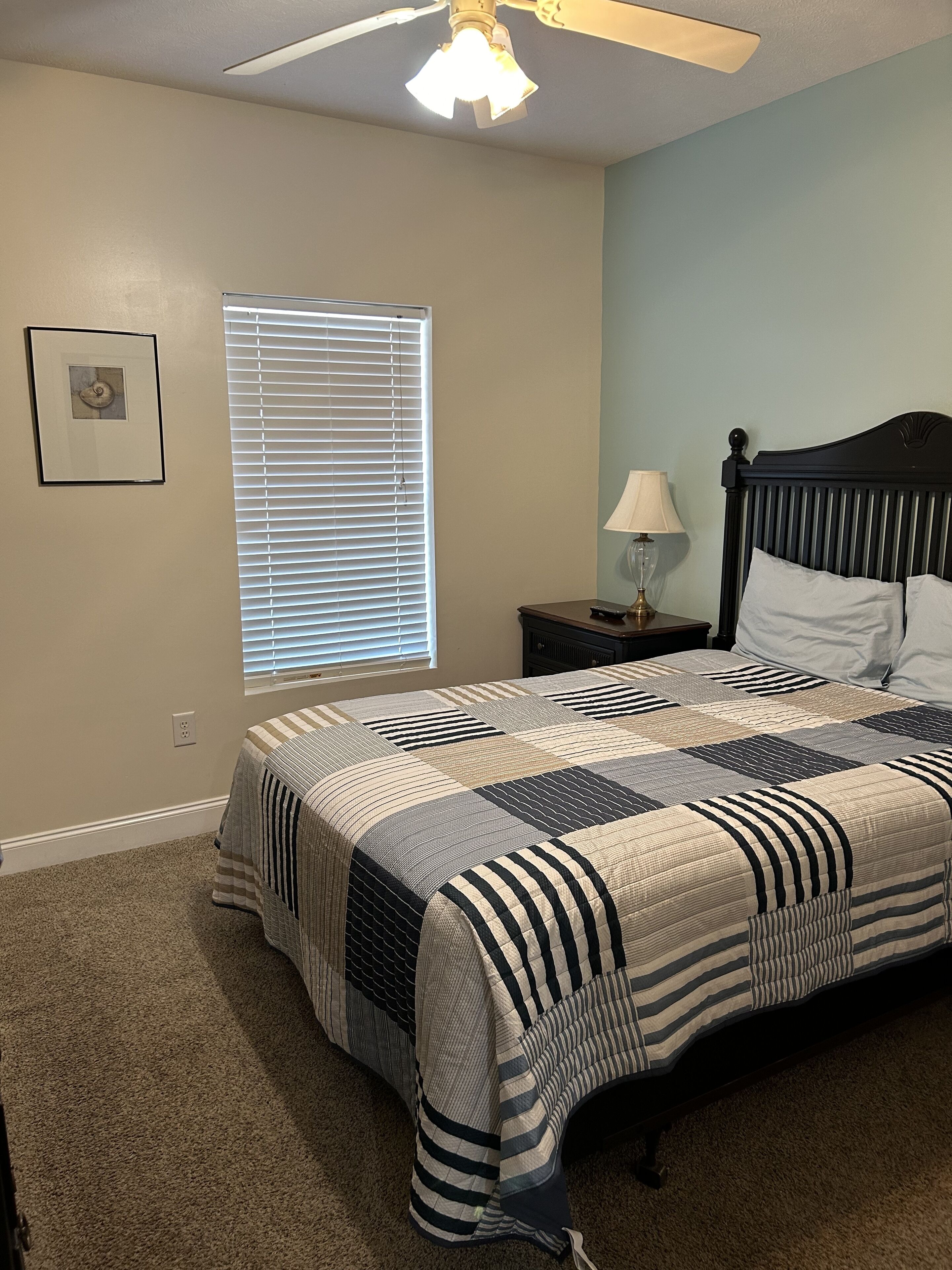 A bedroom with a bed, a nightstand, a lamp, a ceiling fan, a window with blinds, and a framed picture on the wall.