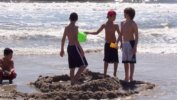 Eine Strandansicht mit spielenden Kindern im Sand und im Hintergrund brechenden Wellen.