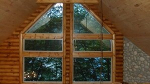 A log cabin interior with a large central window, wooden beams, and a stone wall.