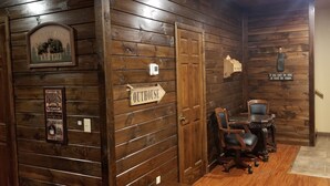 A wooden interior with a sign for 'OUTHOUSE' and a 'WELCOME' sign with a bear and trees. There's a table with chairs and a sign saying 'If you can be kind, at least be vague.'