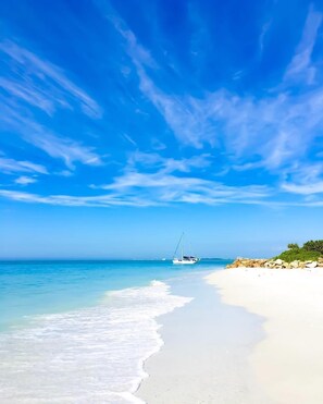 A beach with clear blue water, a white sandy shore, and a sailboat in the distance.