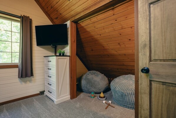 Une pièce avec un plafond en pente, un téléviseur fixé au mur, une commode avec des tiroirs, deux fauteuils-pouf ronds et une porte.