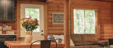 A wooden cabin interior with a dining table, chairs, a sofa, and a sign indicating a 'Lazy Bear Sleeping Bag' is for sale.