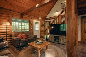Ein Holzblockhaus-Interieur mit Holzbalkendecke, einer Couch, einem Fernseher und einer Treppe.