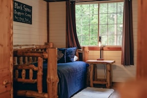A rustic cabin interior with a bed, a small table, a lamp, and a window with curtains.