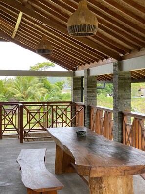 Un espace de repas extérieur en bois, avec un banc et une table, entouré d’une balustrade en bois et offrant une vue sur une végétation luxuriante.