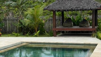 A pool with a thatched-roof gazebo, surrounded by tropical vegetation.