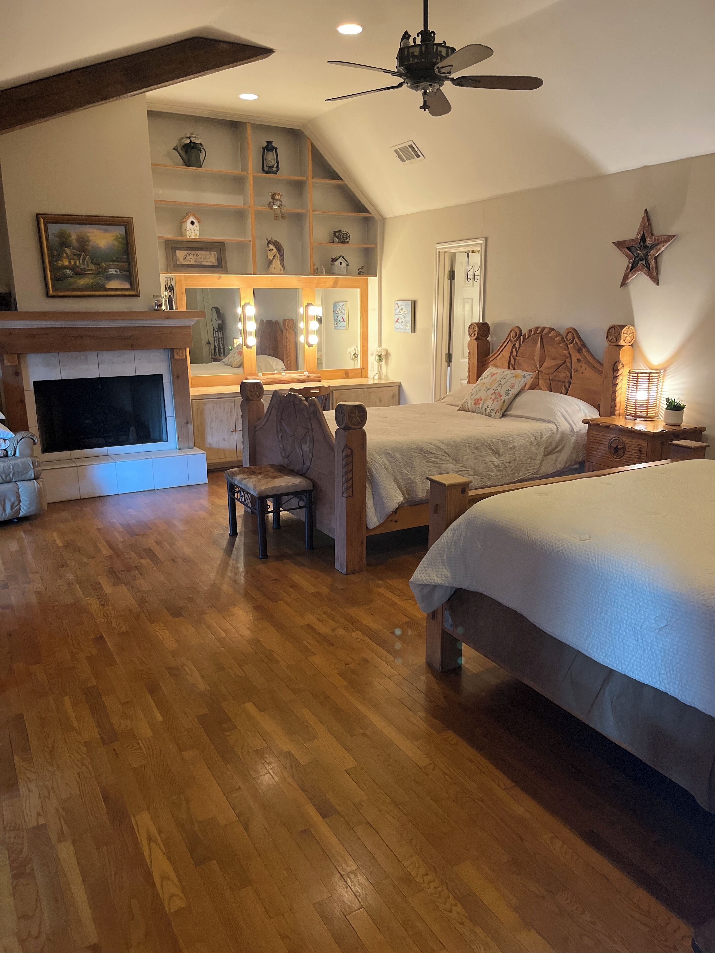 A cozy bedroom with a fireplace, a vanity with mirrors, a bed with a wooden headboard, and a ceiling fan.