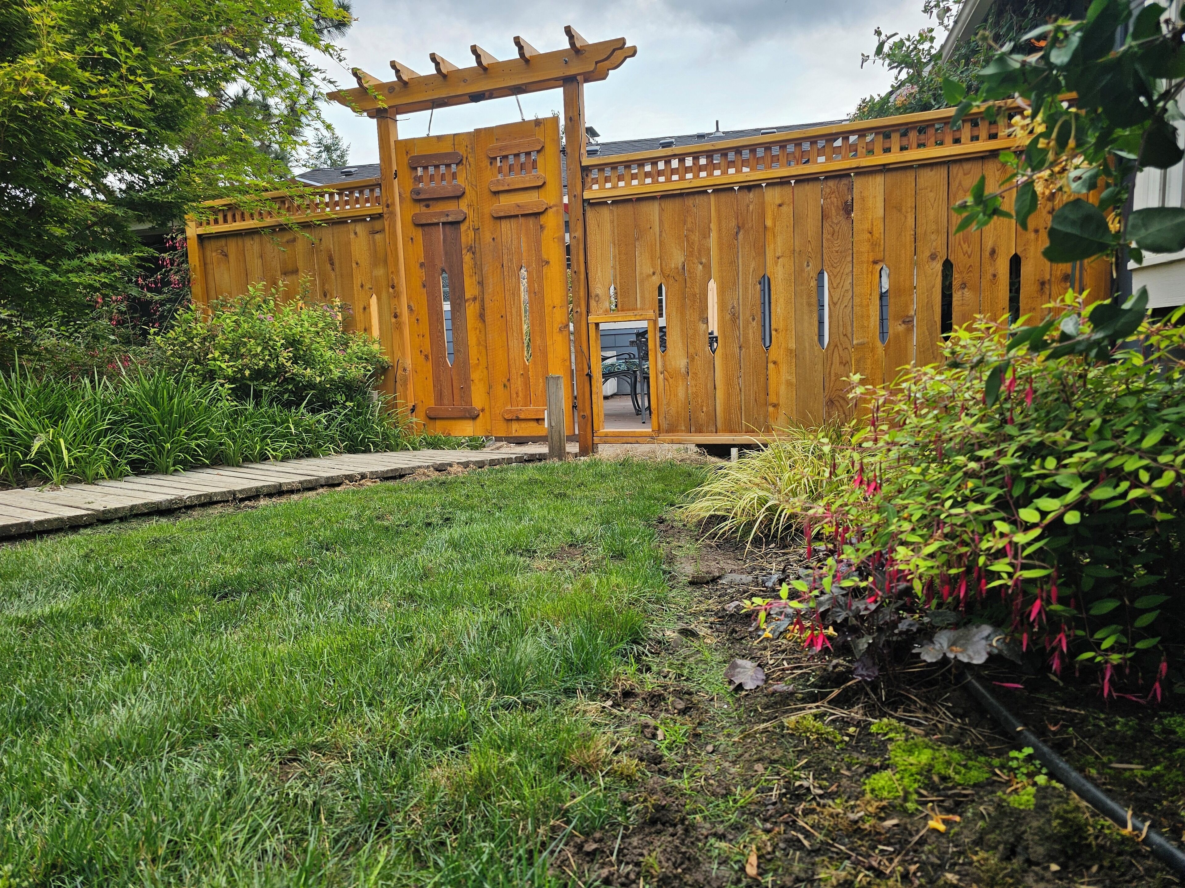 A wooden fence with a pergola entrance, surrounded by a grassy area and various plants.