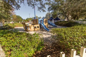 Children can play on the playground's slide, climbing area, and bridge amidst lush greenery and trees.