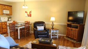 A living room with a brown leather couch, a dining table with chairs, a black leather chair, a television on a wooden stand, and a kitchen area with wooden cabinets and a microwave. - June’s Place In The Country  (Galax)