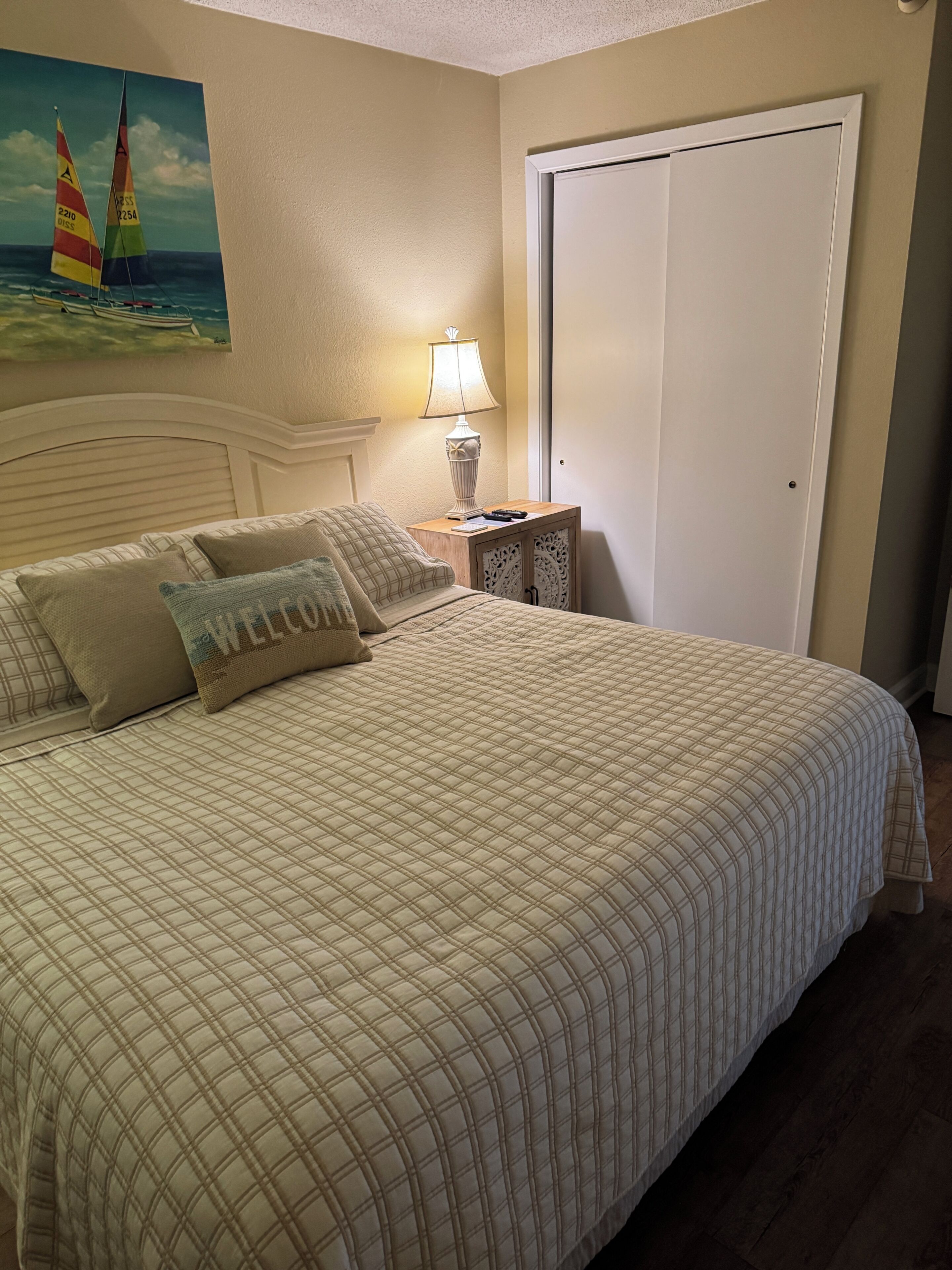 A neatly made bed with a quilted bedspread, a 'WELCOME' pillow, and a painting of sailboats on the wall.