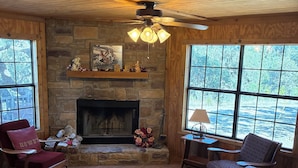 A cozy living room with a stone fireplace, wooden ceiling, and large windows offering a view of the outdoors. - One and a half miles from the Frio River! The stars at night are big and bright! (Leakey)