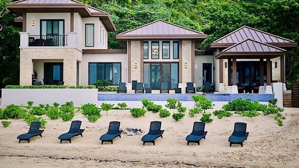 Una casa moderna de dos pisos con balcón, un patio cubierto y una playa con sillas de descanso.