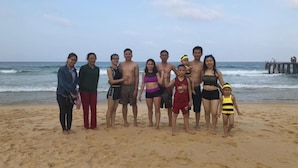 A group of people standing on a sandy beach with the ocean and a pier in the background. - Anh Quan Hotel (Quy Nhon Nam)