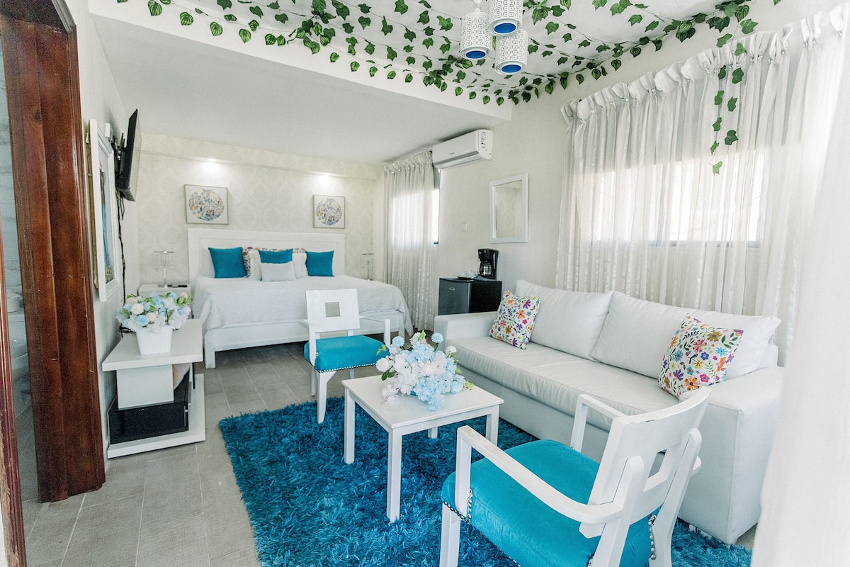 A modern, well-lit room with a bed, white furniture, a coffee table, and a blue rug.