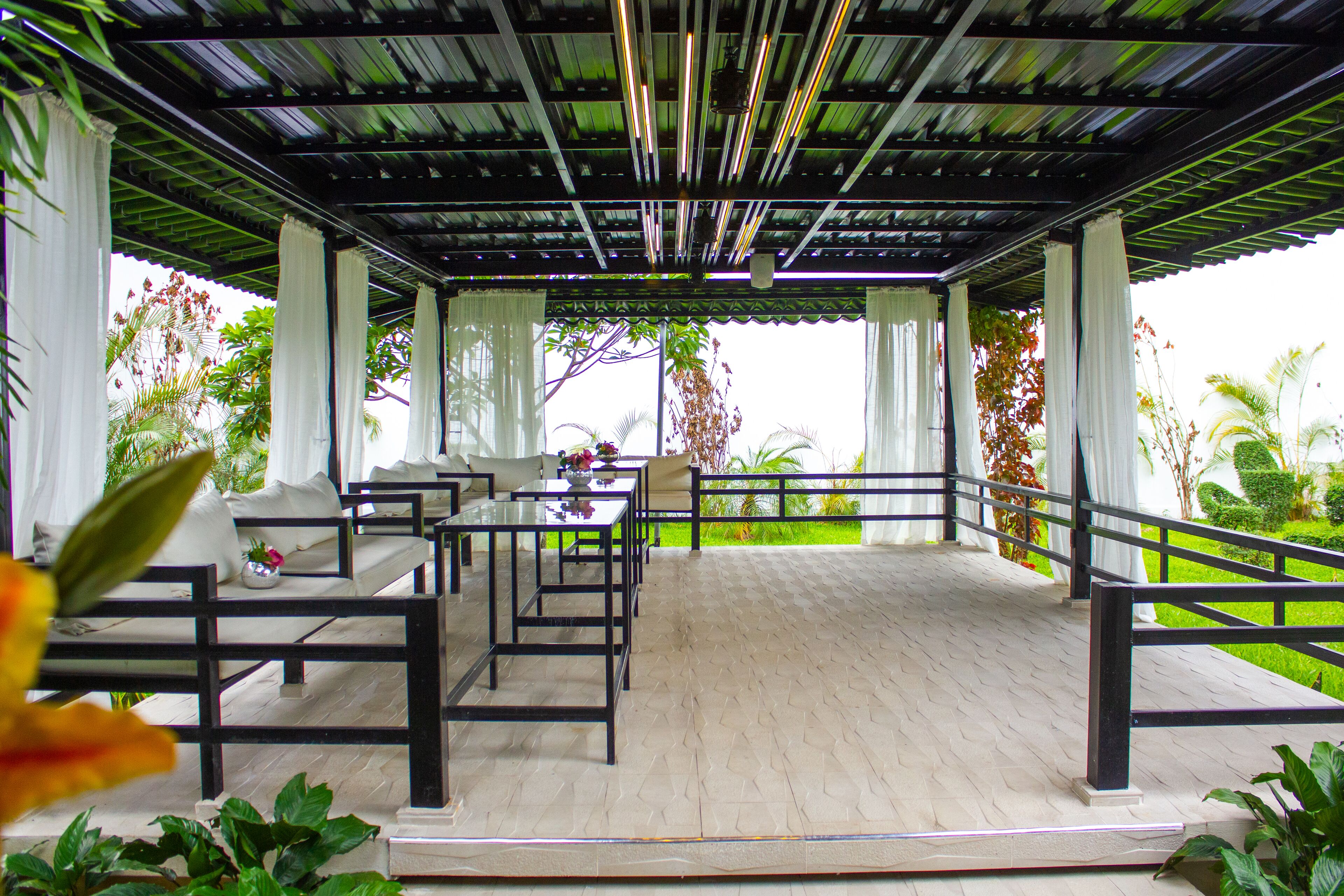 A covered outdoor seating area with tables and chairs, surrounded by greenery and a view of the landscape.