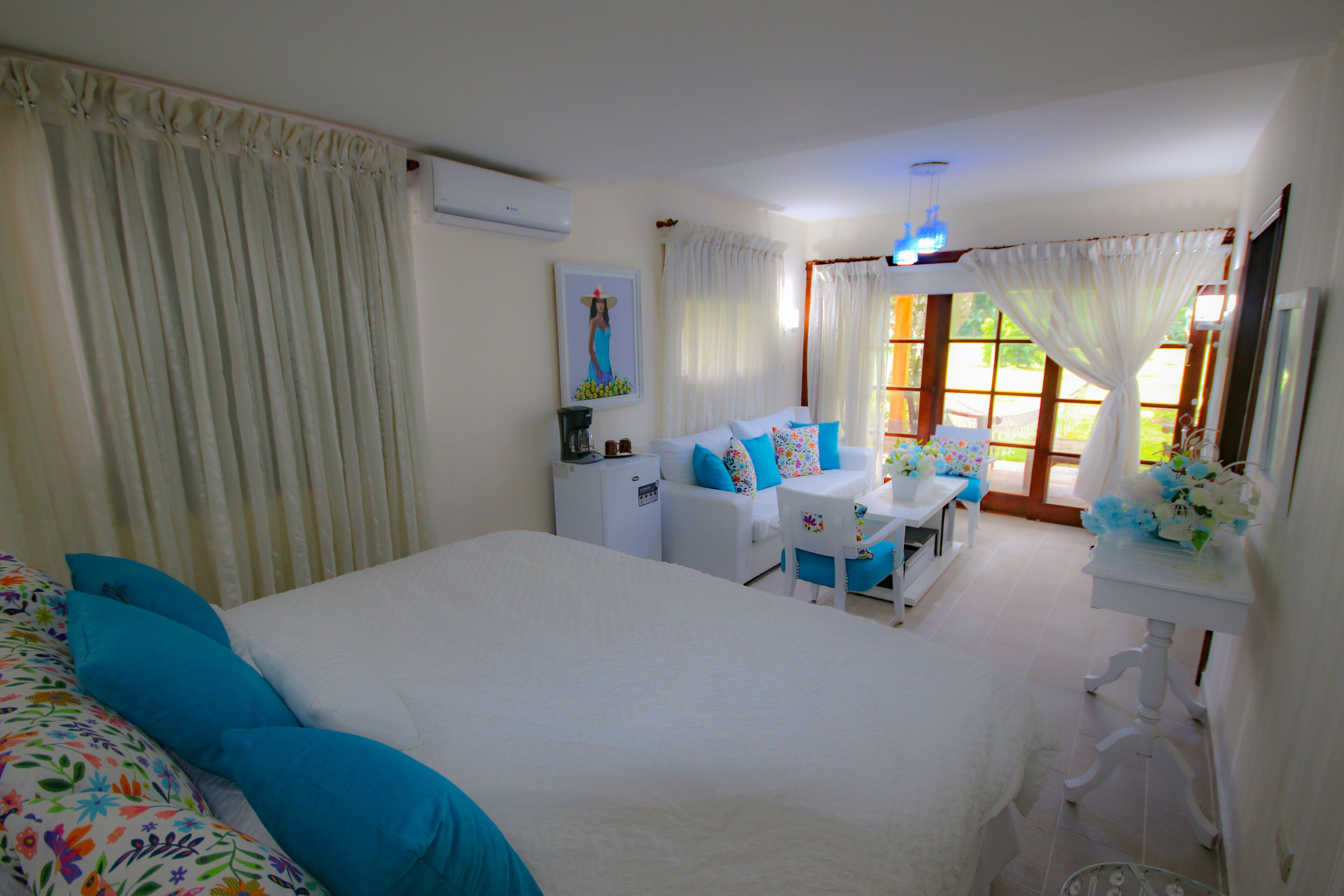 A bedroom with a bed, a desk, and a chair. There is a window with curtains, a small table with a vase of flowers, and a wall-mounted air conditioner.