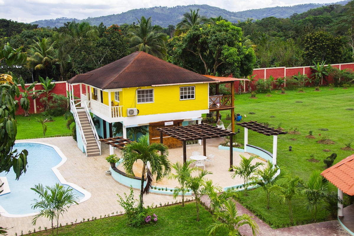 A yellow house with a swimming pool, surrounded by greenery and a red wall.