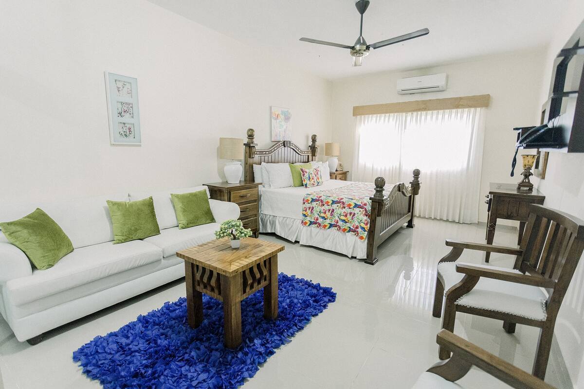 A well-lit, modern bedroom with a white bed, green pillows, a wooden nightstand, a blue rug, and a ceiling fan.