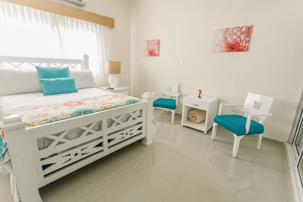 A bedroom with a bed, two chairs, a nightstand, and a wall-mounted shelf.