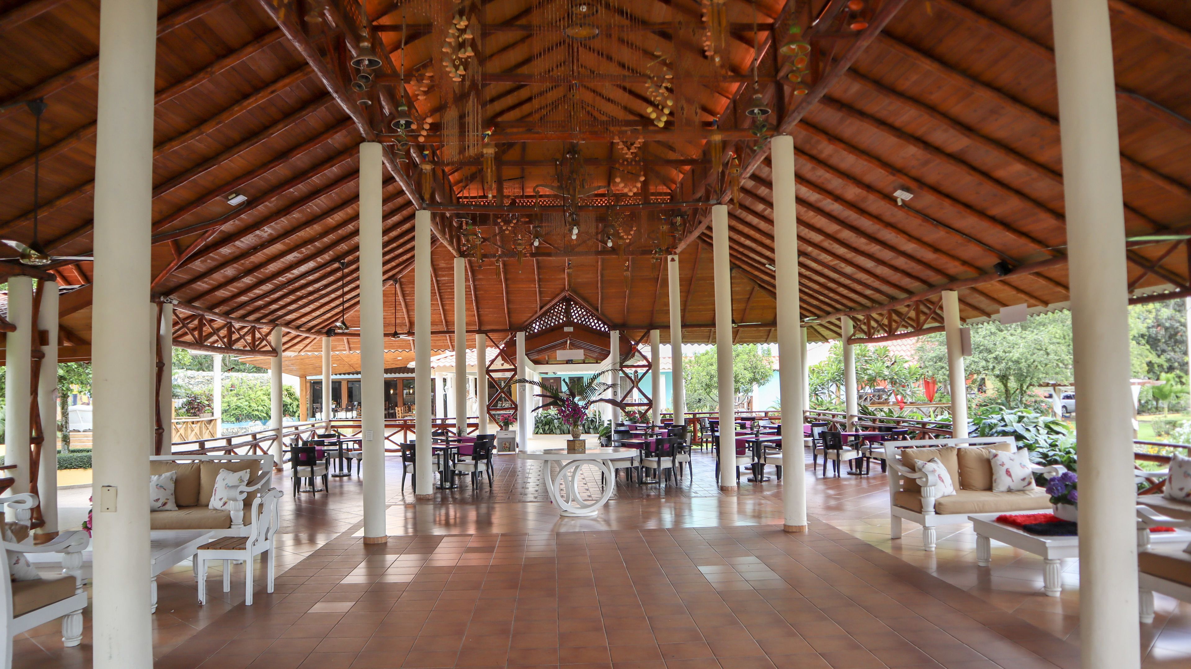 A covered outdoor seating area with white columns, tables, and chairs, featuring a central decorative element and a view of greenery outside.