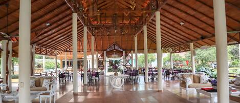 A covered outdoor seating area with white columns, tables, and chairs, featuring a central decorative element and a view of greenery outside.
