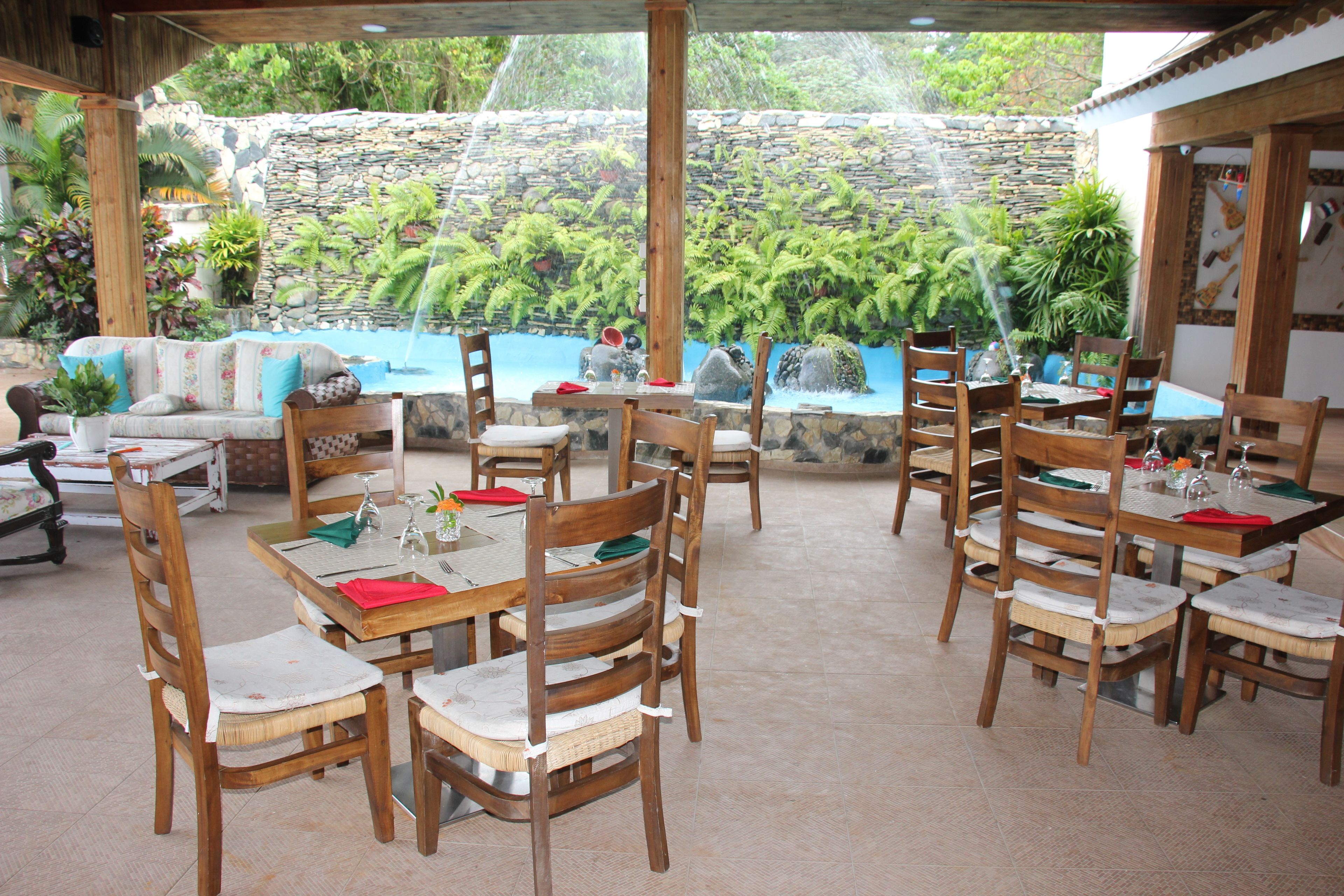 An outdoor dining area with wooden tables and chairs, a stone wall, and lush greenery in the background.