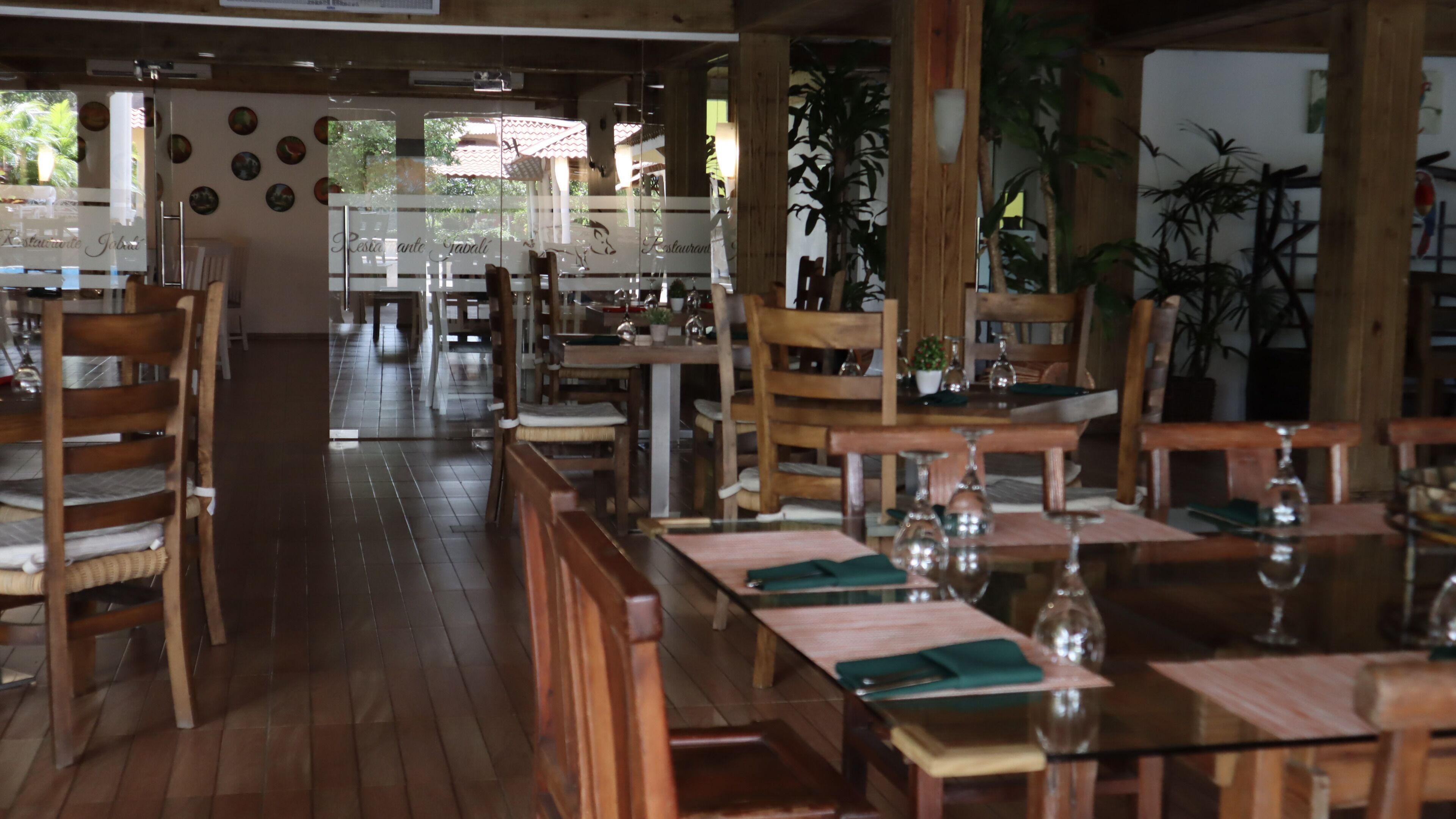 Un restaurante con mobiliario de madera, mesas de vidrio y plantas en macetas.