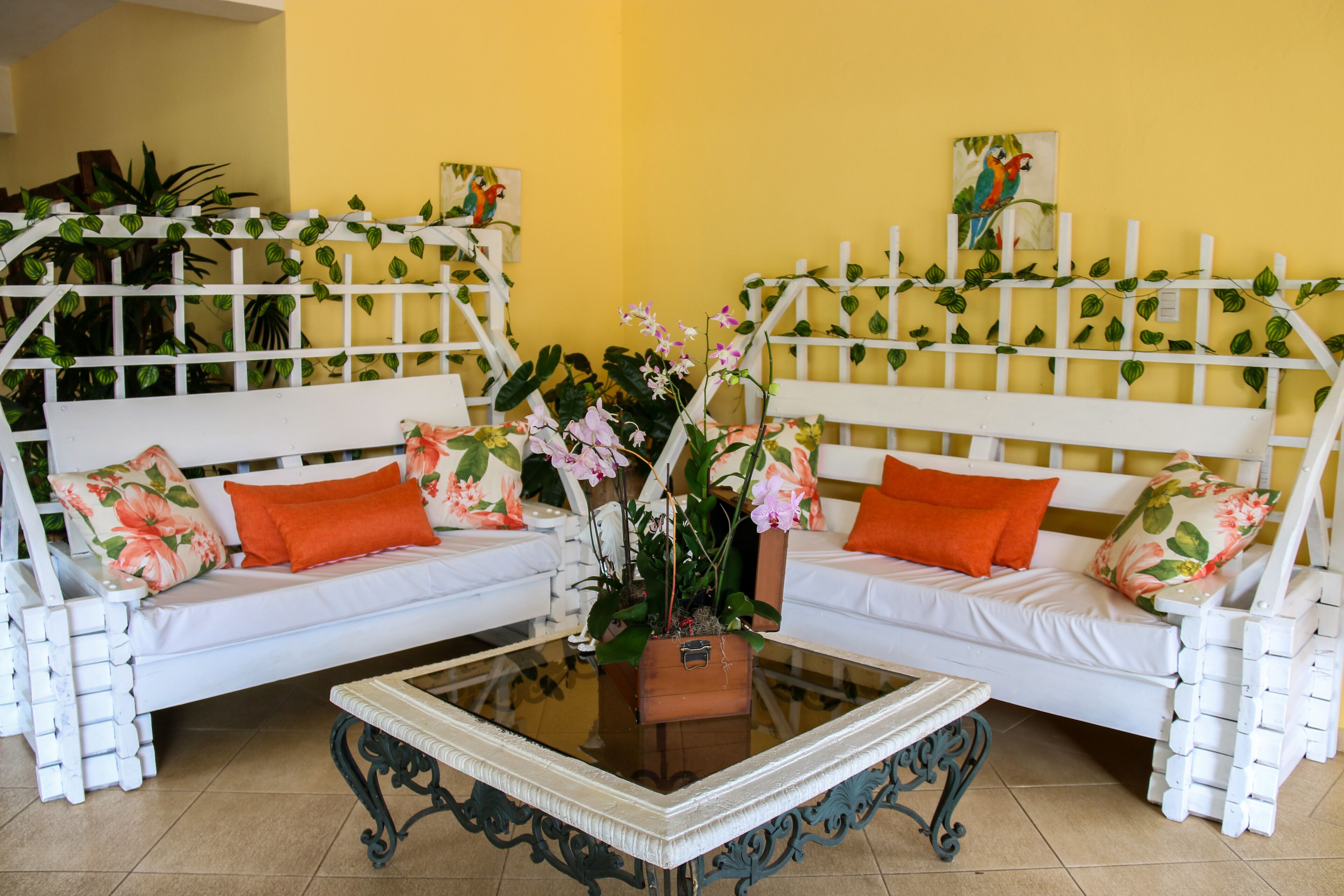 Una habitación con dos sofás blancos con cojines naranjas, una mesa de centro de vidrio y una cerca blanca con plantas.