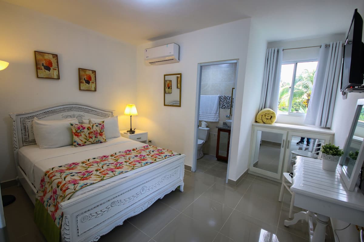 A bedroom with a bed, bedside table, lamp, air conditioning unit, and a window with curtains.