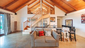 A spacious living area with a high ceiling, a staircase leading to an upper level, a modern kitchen, and a seating area with a sofa and chairs. - Outlook Hill Vineyard Cottages (Tarrawarra)