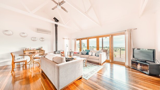 A spacious living room with a sofa, a dining table, and a TV. There is a large window with a view of the outdoors. - Outlook Hill Vineyard Cottages (Tarrawarra)