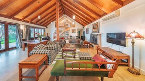 A spacious living area with a high wooden ceiling, a staircase leading to an upper level, a large TV, and a dining area with a table and chairs. - Outlook Hill Vineyard Cottages (Tarrawarra)