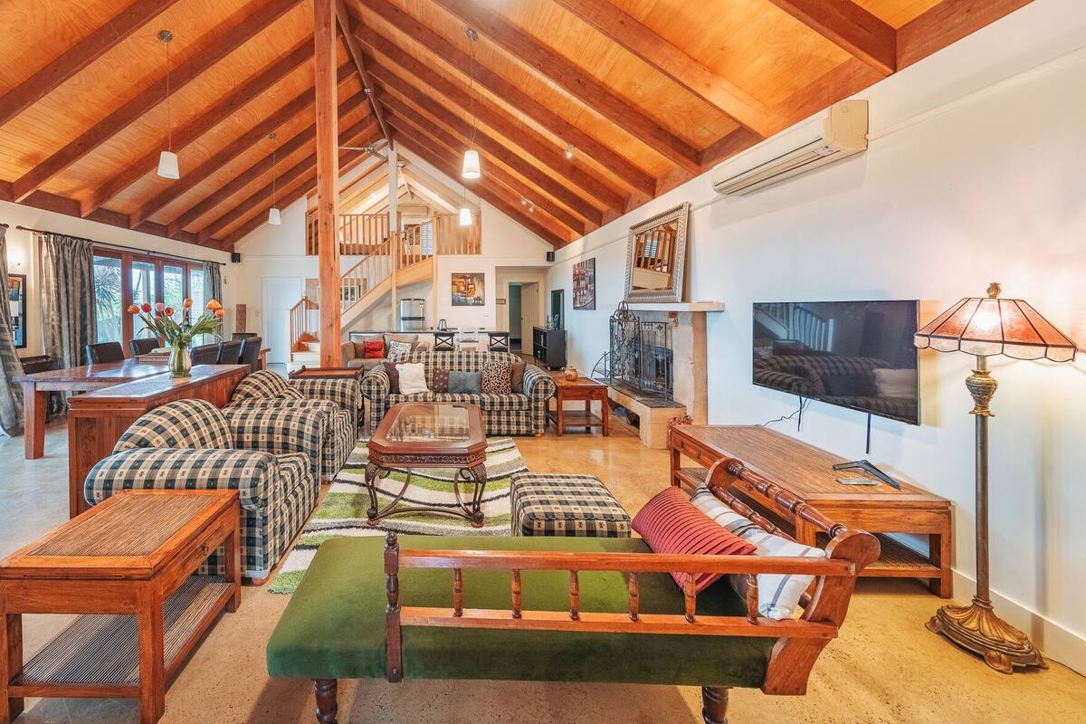 A spacious living area with a high wooden ceiling, a staircase leading to an upper level, a large TV, and a dining area with a table and chairs.