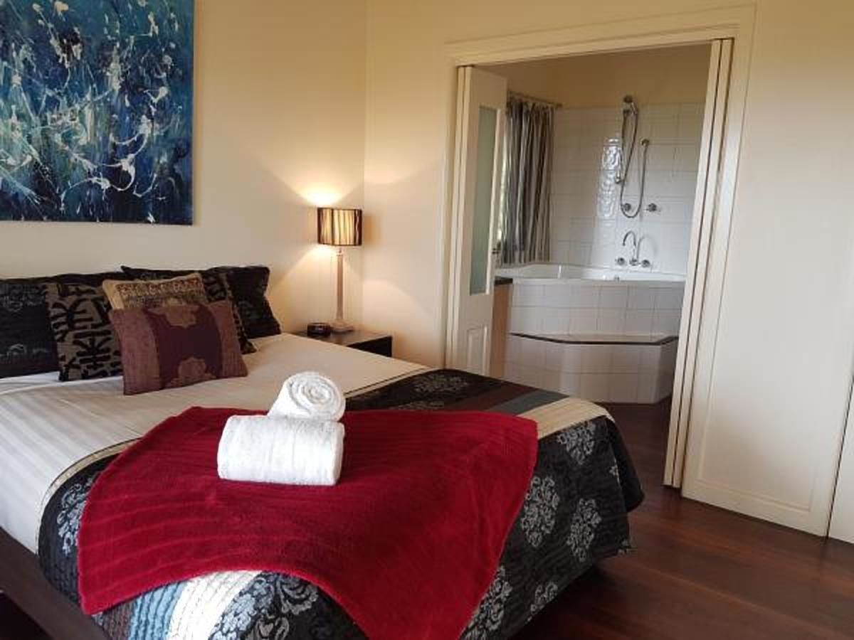 A hotel room with a bed, a red throw blanket, a folded towel, a nightstand, a lamp, a painting on the wall, and a bathroom with a shower and sink.