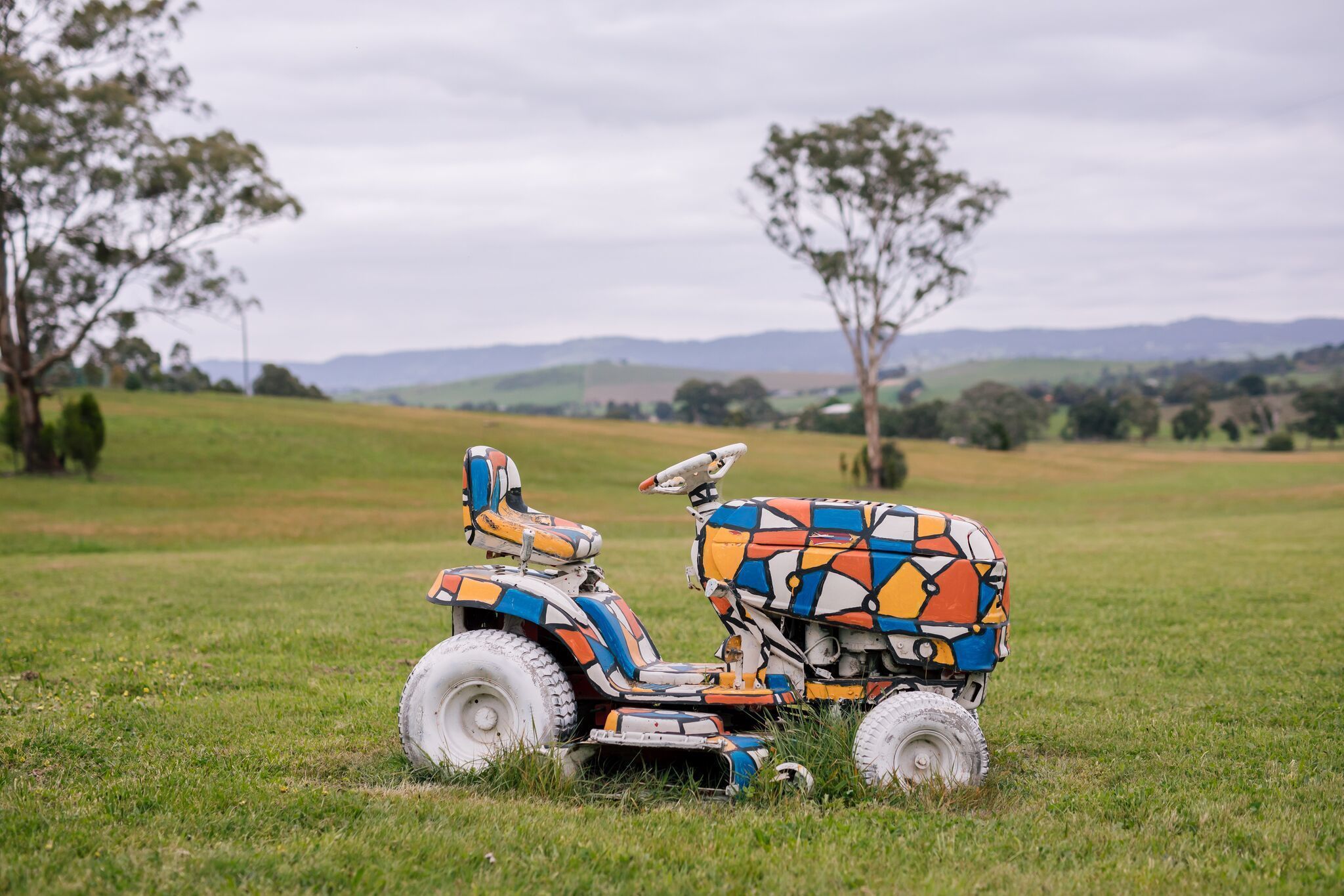 A colorful lawnmower in a grassy field with trees and hills in the background.