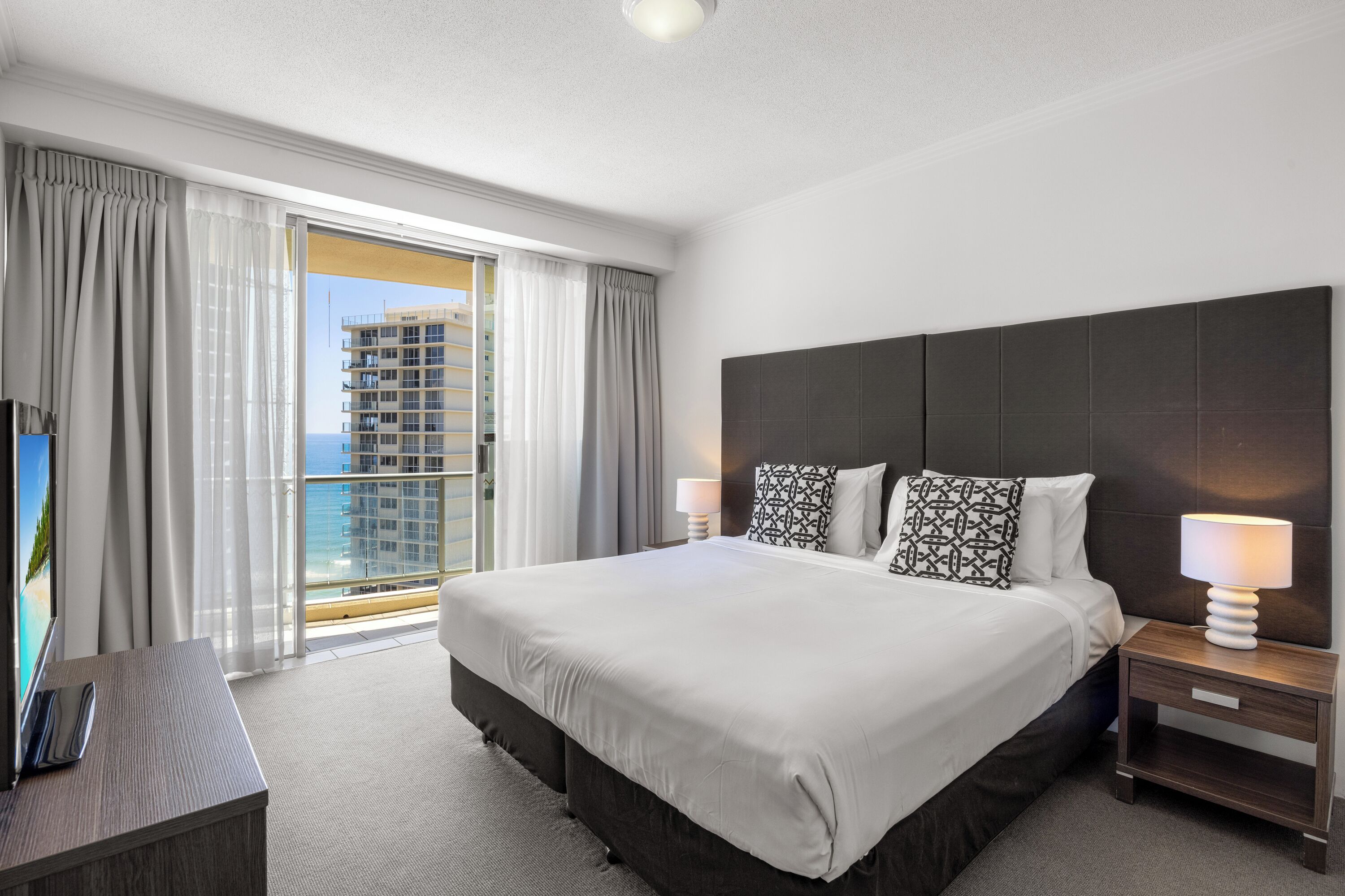 A hotel room with a large bed, two bedside tables with lamps, a TV on a stand, and a balcony with curtains.