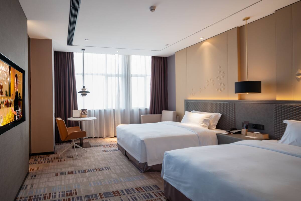 A hotel room with two beds, a small table, a chair, and a TV on the wall.