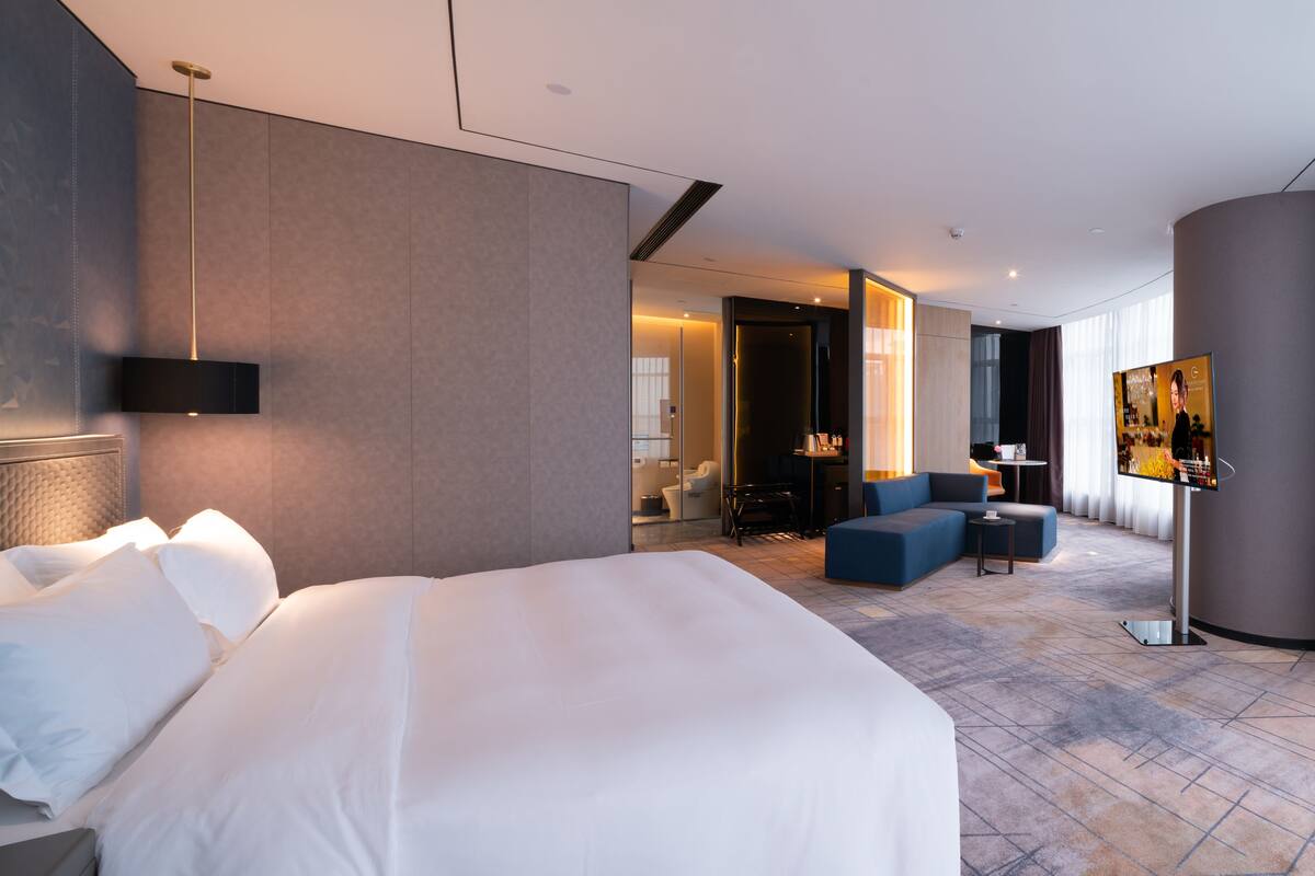 A modern hotel room with a large bed, a seating area with a sofa and armchairs, a flat-screen TV, and a glass partition separating the room from the bathroom.