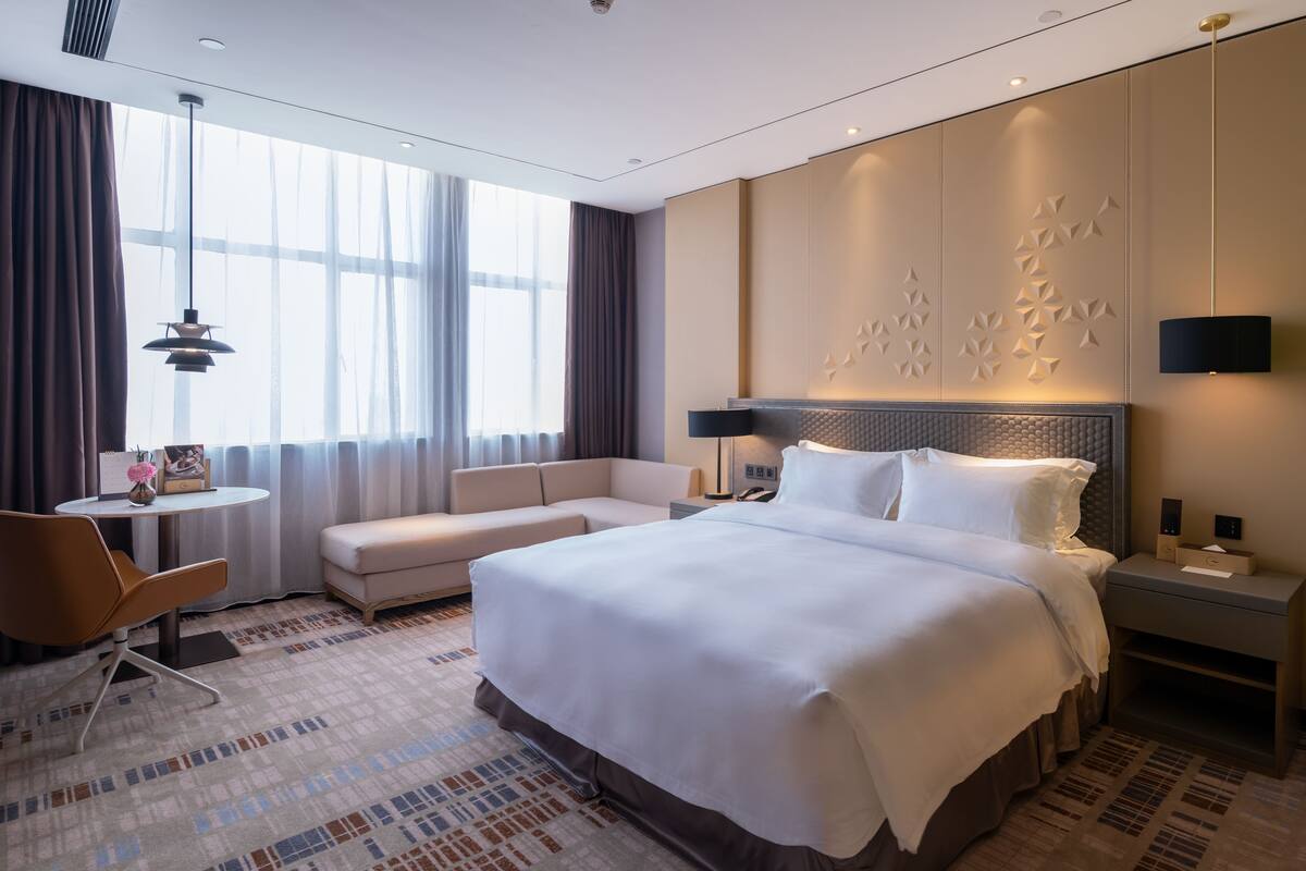 A modern hotel room with a large bed, a sofa, a desk, and a large window.