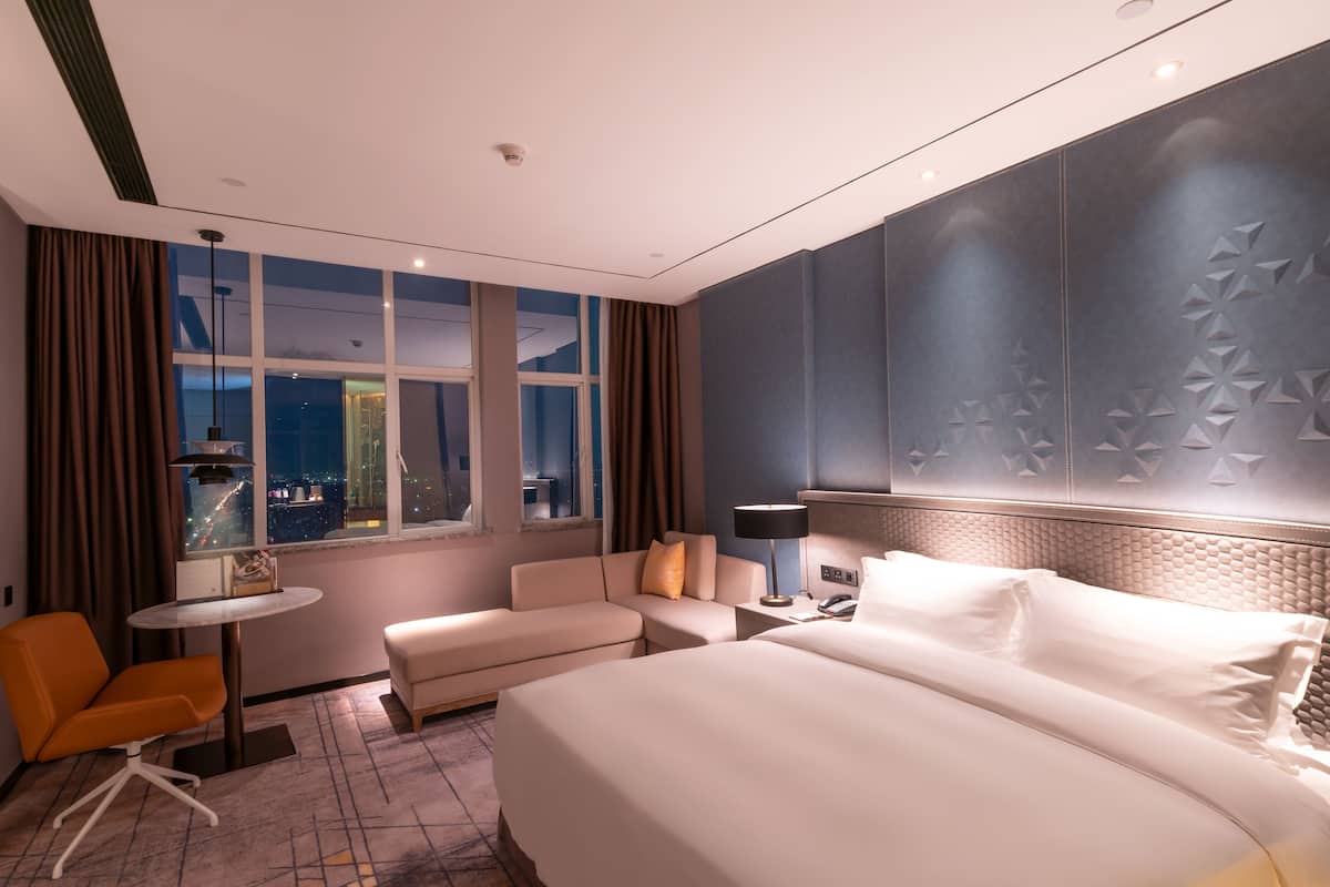 A modern hotel room with a large bed, a sofa, a small table, and a view of the city at night.